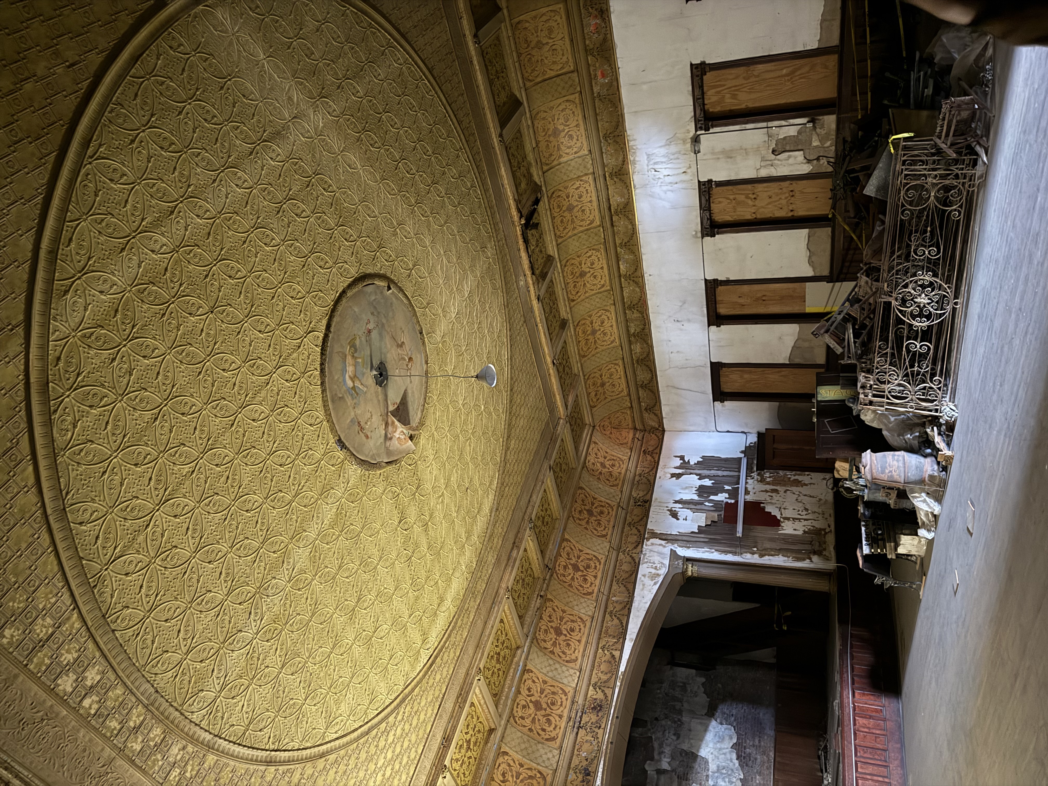 Yellow, ornate ceiling with a single hanging bulb. A stage is in the background and old materials are against the side of the room, natural light coming through and open door.