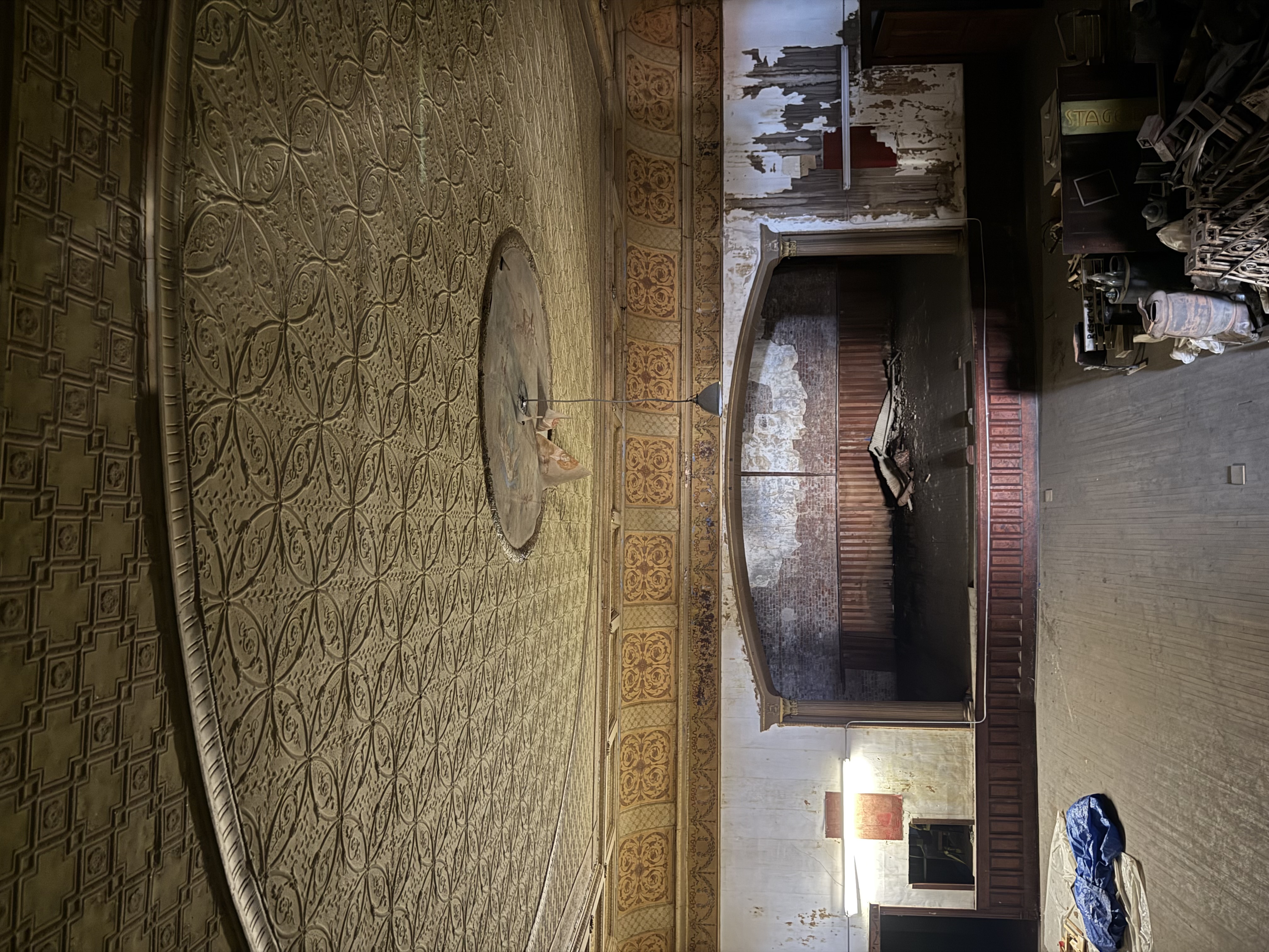 A yellowed, ornate ceiling with decorative moulding in vivid light shows wear. A single lamp hangs with torn paper from the ceiling.