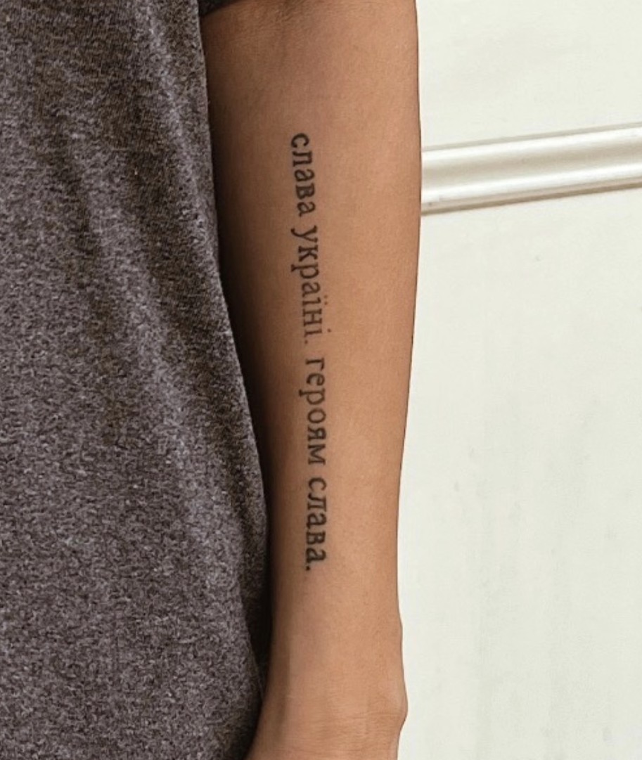 A partial view of a dark grey t-shirt with a tattoo on a forearm reading A partial view of a dark grey t-shirt with a tattoo on a forearm reading “Слава Україні. Героям слава” “Glory to Ukraine. Glory to Heroes.”